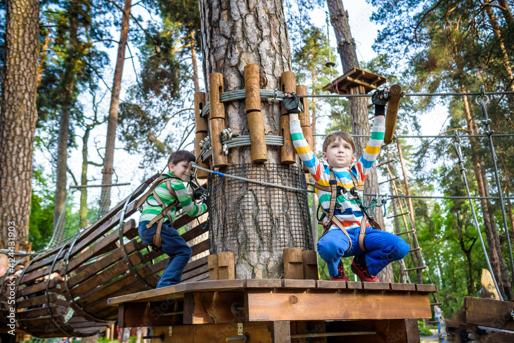 Two little boy boys kid in a helmet and with a safety carbine goes on a rope on the background of forest and sky. Healthy games outdoor