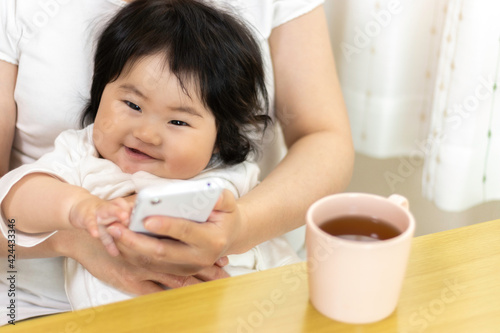 赤ちゃんを膝に座らせて、スマートフォンを使う女性。赤ちゃん（0歳、生後8か月、日本人、女の子）