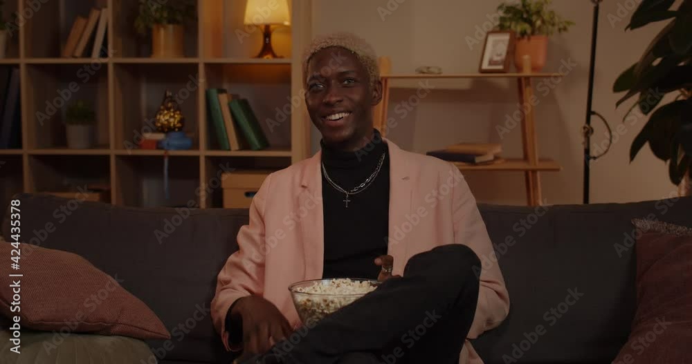 Crop view of millennial cheerful man taking remote controller and switching on tv. Afro american male person holding bowl with popcorn while sitting comfortably on couch and watching film. Crop view of millennial cheerful man taking remote controller and switching on tv. Afro american male person holding bowl with popcorn while sitting comfortably on couch and watching film.