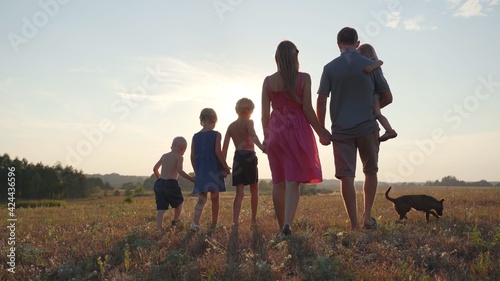 A friendly large family walks across the field at sunset with dog.