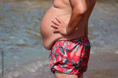 Overweight, obesity and all inclusive concept. Man in trunks with fat belly standing on a beach photo