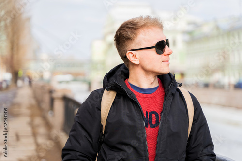 A young man uses a mobile phone and stands on the embankment of St. Petersburg with a view of old buildings, early spring