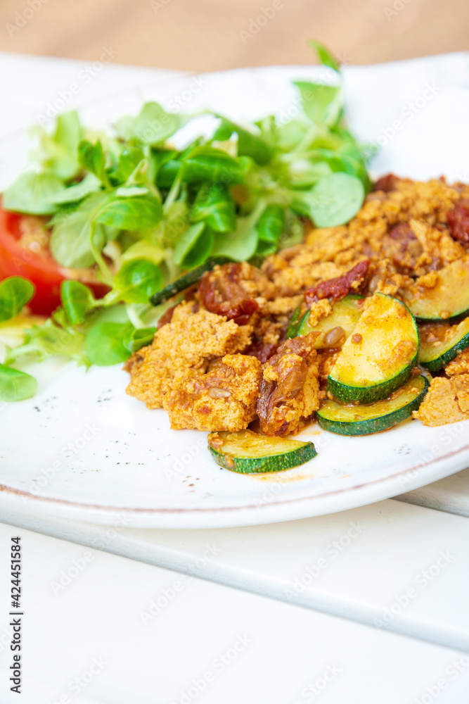 Vegan breakfast prepared with scrambled tofu and eggs, cooked with dried tomatoes and zucchini served with fresh lettuce and cucumber in white ceramic vintage plate