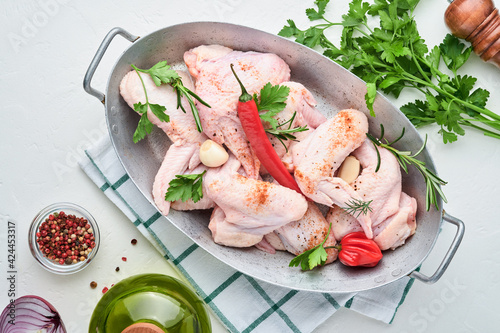 Raw chicken wings in a white bowl with spices and ingredients for cooking on light slate, stone or concrete background. Top view with copy space. Mock up.