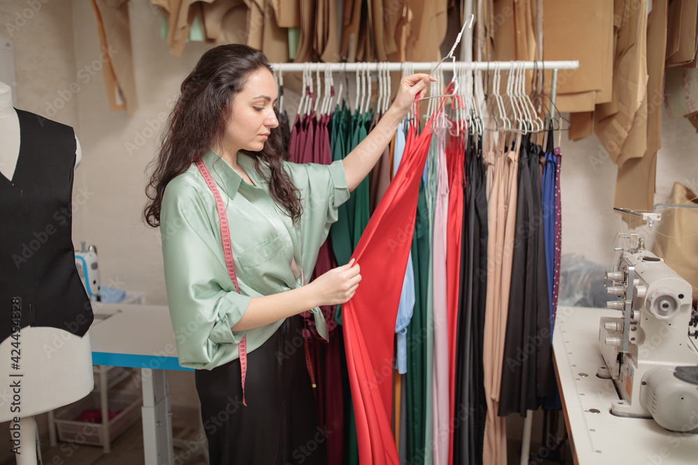 Female fashion designer working on new womenswear collection for clients in cozy workshop studio, dressmaker, tailor or needlewoman standing near clothing rack with fashionable stylish clothes