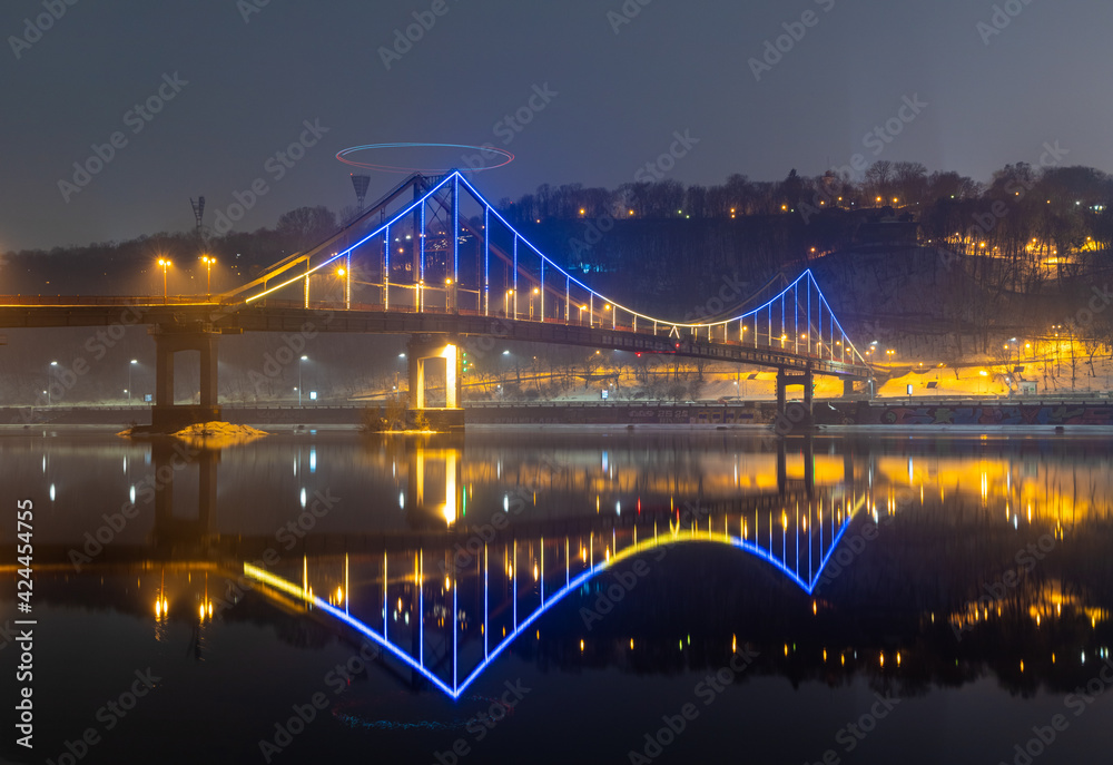 Pedestrian bridge in Kiev. Evening lighting. Reflection of the bridge in the river. Pedestrian bridge in Kiev. Evening lighting. Reflection of the bridge in the river.
