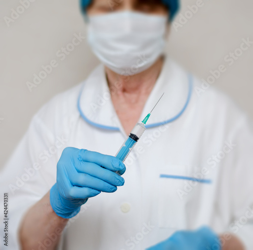 A woman's hand in blue medical glove on the background of white coat holds syringe, close-up.