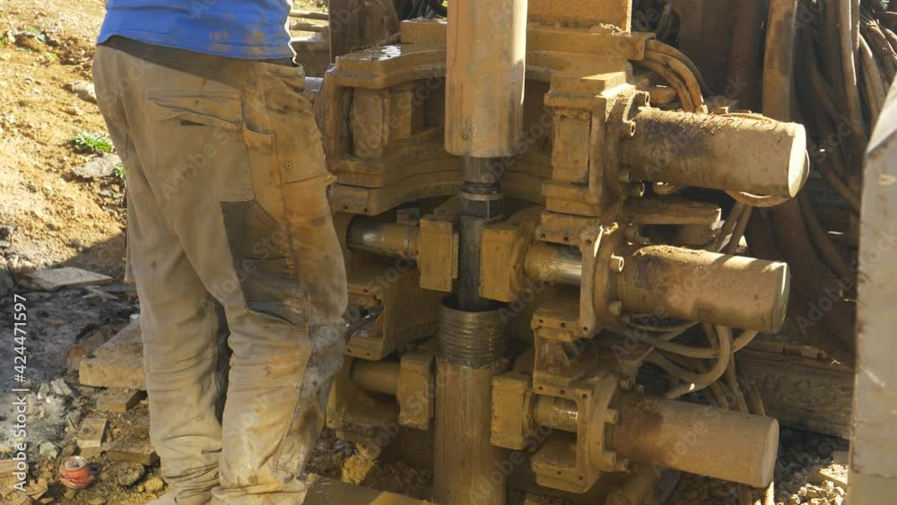 CLOSE UP: Unrecognizable male contractor stands next to a drill boring a hole in the ground near a construction site. Worker oversees the boring process during modern geothermal pump installation. CLOSE UP: Unrecognizable male contractor stands next to a drill boring a hole in the ground near a construction site. Worker oversees the boring process during modern geothermal pump installation.