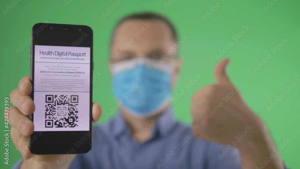 Young man with face mask shows smartphone for scanning the digital health certificate, thumb up OK sign to encourage others to use it, front view isolated on green screen chroma key Young man with face mask shows smartphone for scanning the digital health certificate, thumb up OK sign to encourage others to use it, front view isolated on green screen chroma key