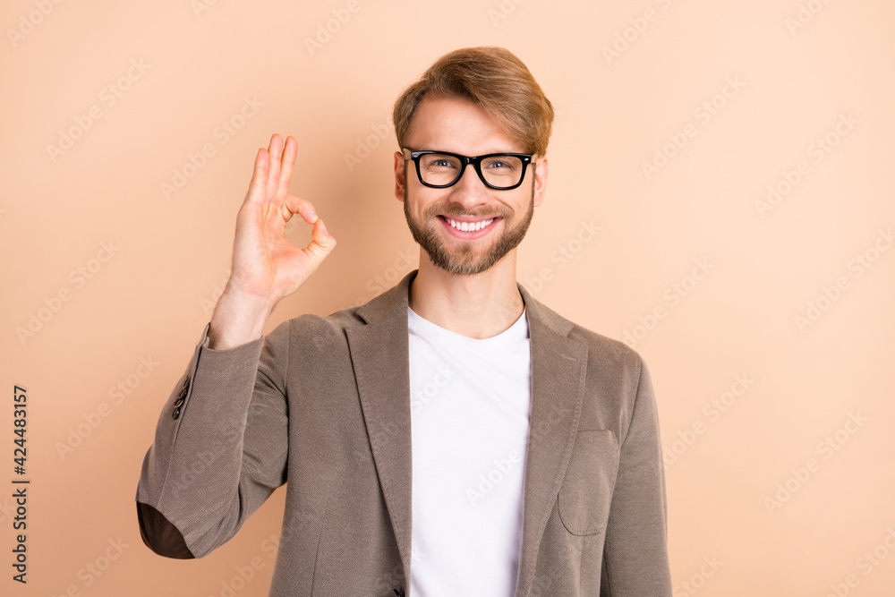 Photo of optimistic nice blond hair man show okey wear spectacles grey jacket isolated on beige background Photo of optimistic nice blond hair man show okey wear spectacles grey jacket isolated on beige background