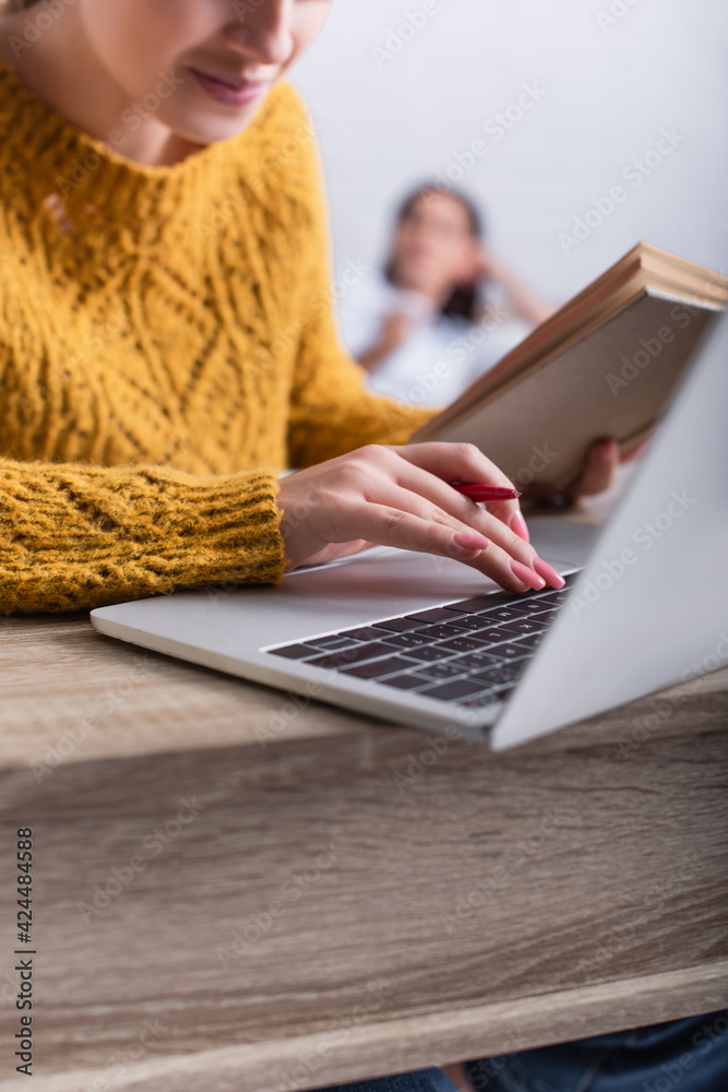 Fototapeta premium cropped view of teenage girl typing on laptop keyboard and holding book
