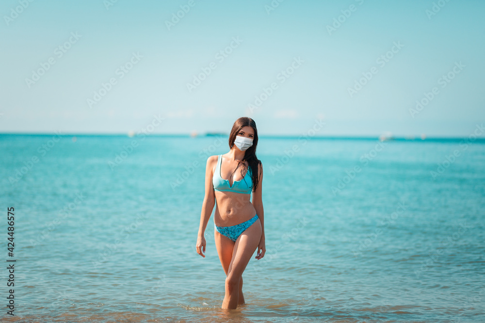 Summer. A young pretty woman wearing a protective mask poses standing at the ocean. The concept of summer holidays during the coronavirus pandemic