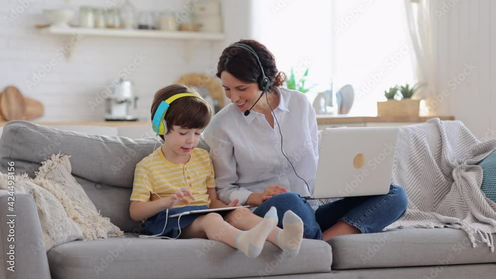 Happy mother with kid sitting on couch at home office during lockdown work on laptop. Child playing in game at tablet sit on sofa, son showing something on tablet to mom. Family, e-learning concept Happy mother with kid sitting on couch at home office during lockdown work on laptop. Child playing in game at tablet sit on sofa, son showing something on tablet to mom. Family, e-learning concept