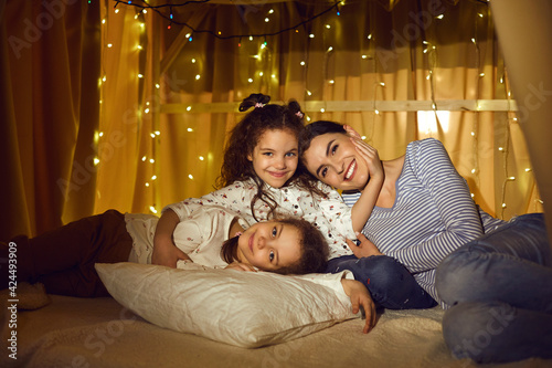 Happy family portrait. Affectionate mother and two little daughter relaxing in tent with flashlights in home living room. Smiling positive caucasian mom hugging with children looking at camera