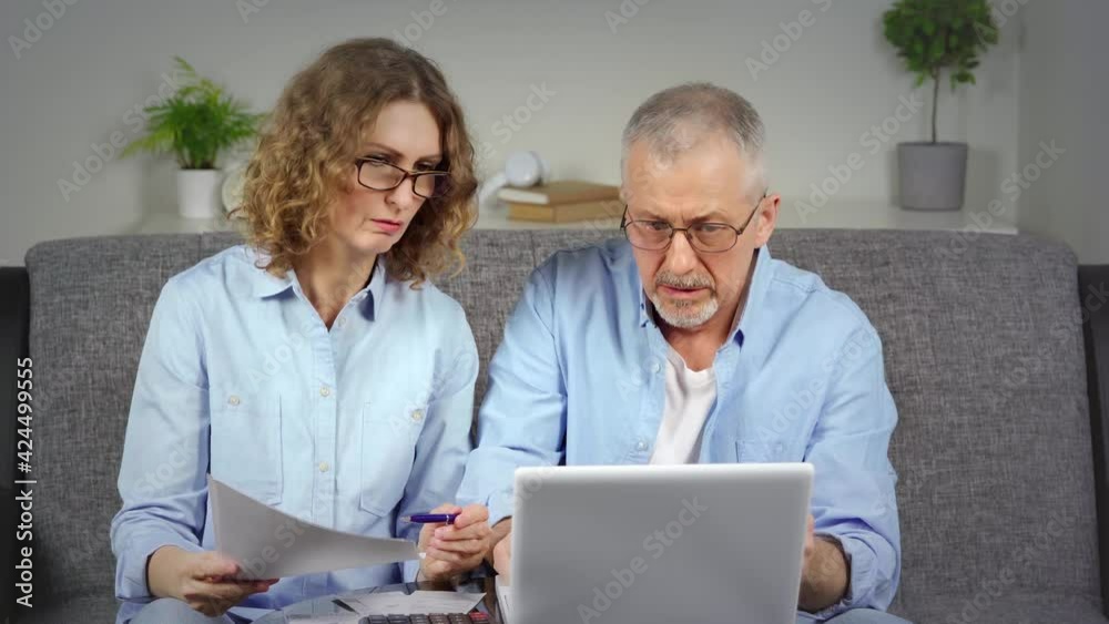 A focused middle-aged woman holds documents in her hands and discusses information with her husband on a laptop. A focused middle-aged woman holds documents in her hands and discusses information with her husband on a laptop.