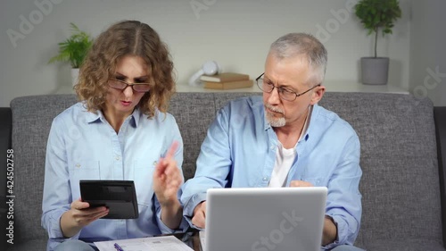 A focused middle-aged woman holds a calculator in her hands and counts the family budget with her husband on a laptop.