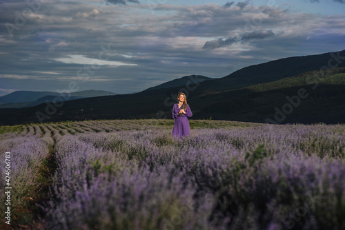 a blonde woman stands in a purple dress in the mountains on a lavender field in the rays of sunlight