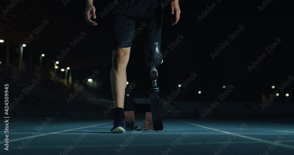 Cinematic close up of disable man with legs prosthesis is warming up before run with dedication on car track at night. Concept of handicapped people active lifestyle, determination, motivation.
