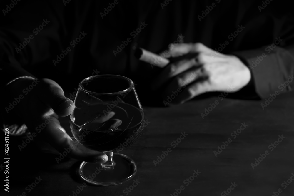 Man hands with a glass of whiskey and a cigar on dark table.Black and white photography,smoke and drink alcohol addiction concept. Man hands with a glass of whiskey and a cigar on dark table.Black and white photography,smoke and drink alcohol addiction concept.