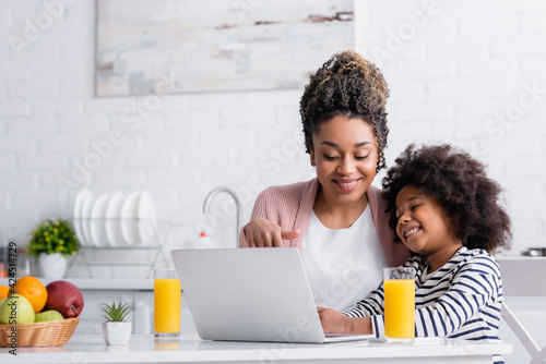 smiling african american woman pointing at laptop while watching movie with daughter in kitchen