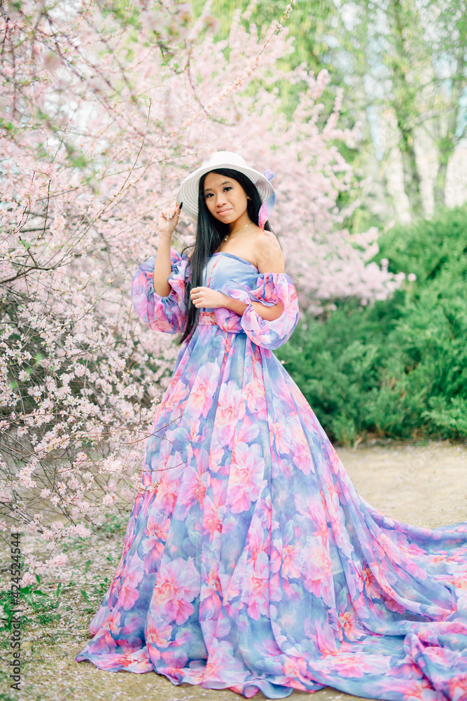 Beautiful young asian brunette woman in luxury long dress among the blossoming sakura flowers. enjoying spring Early spring concept. Rest on nature vacation. Girl in straw hat.