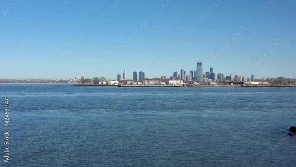 A drone view from Valentino Pier in Brooklyn on a sunny day with blue skies. The camera boom up in front of a fence at the end of the pier. Jersey City is in the distance. A drone view from Valentino Pier in Brooklyn on a sunny day with blue skies. The camera boom up in front of a fence at the end of the pier. Jersey City is in the distance.