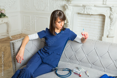 Brown-hair woman wearing blue medical scrubs sits on a sofa with syringe in her hand photo