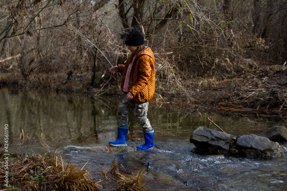 The girl in rubber boots in a brown jacket crosses the river ford, in early spring the trees around without leaves. Fun childhood The girl in rubber boots in a brown jacket crosses the river ford, in early spring the trees around without leaves. Fun childhood
