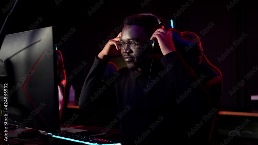 An african student sits in a gaming chair in a computer club, puts on headphones, smiles, and prepares for the game