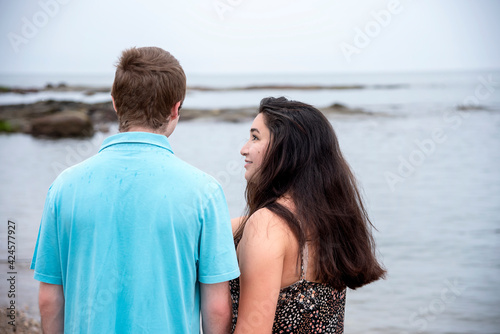 Casual outdoor back view portrait of a cute young couple in love walking close together on the beach in Guilford, CT with the woman's face in profile as she looks at the man  