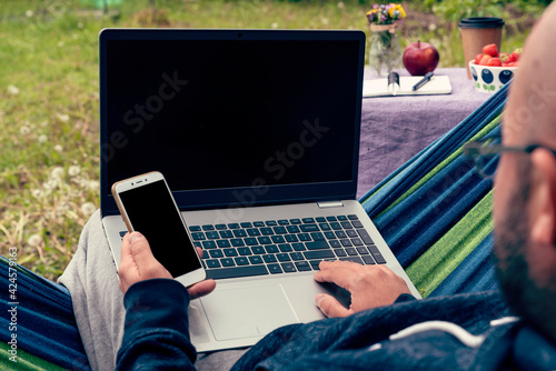 Man typing on laptop and holding a mobile in hand sitting on hammock outdoors during home office. Work as a freelancer with internet and keyboard. Blank minitor with mock up.  photo