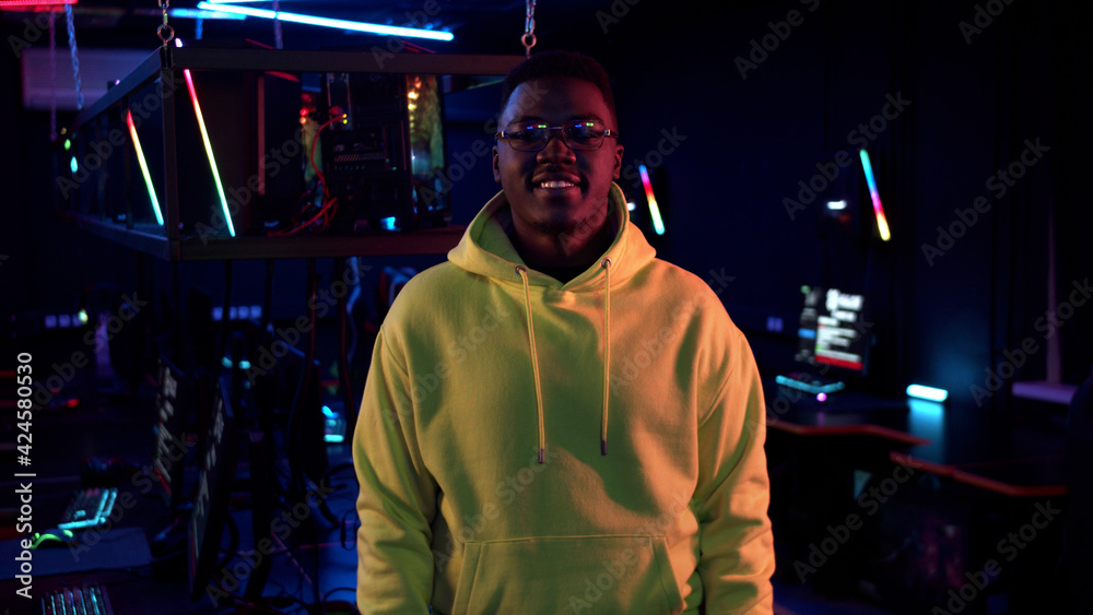 Portrait of a young smiling black guy in a yellow hoodie against the background of a computer room in neon light Portrait of a young smiling black guy in a yellow hoodie against the background of a computer room in neon light