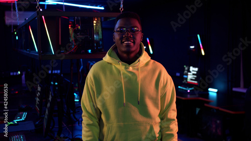 Portrait of a young smiling black guy in a yellow hoodie against the background of a computer room in neon light