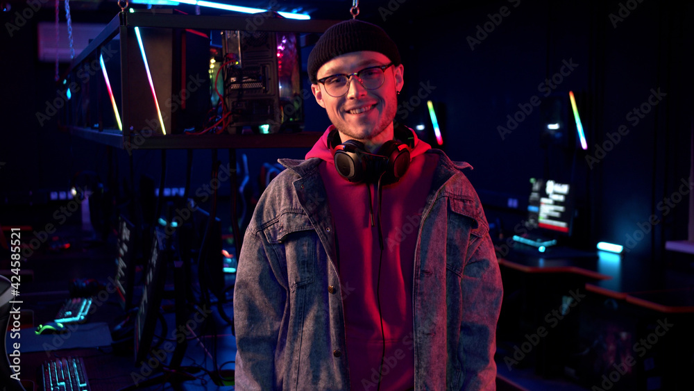 In a computer club in neon light, a young guy in a hat, glasses and a denim jacket poses for the camera and smiles In a computer club in neon light, a young guy in a hat, glasses and a denim jacket poses for the camera and smiles