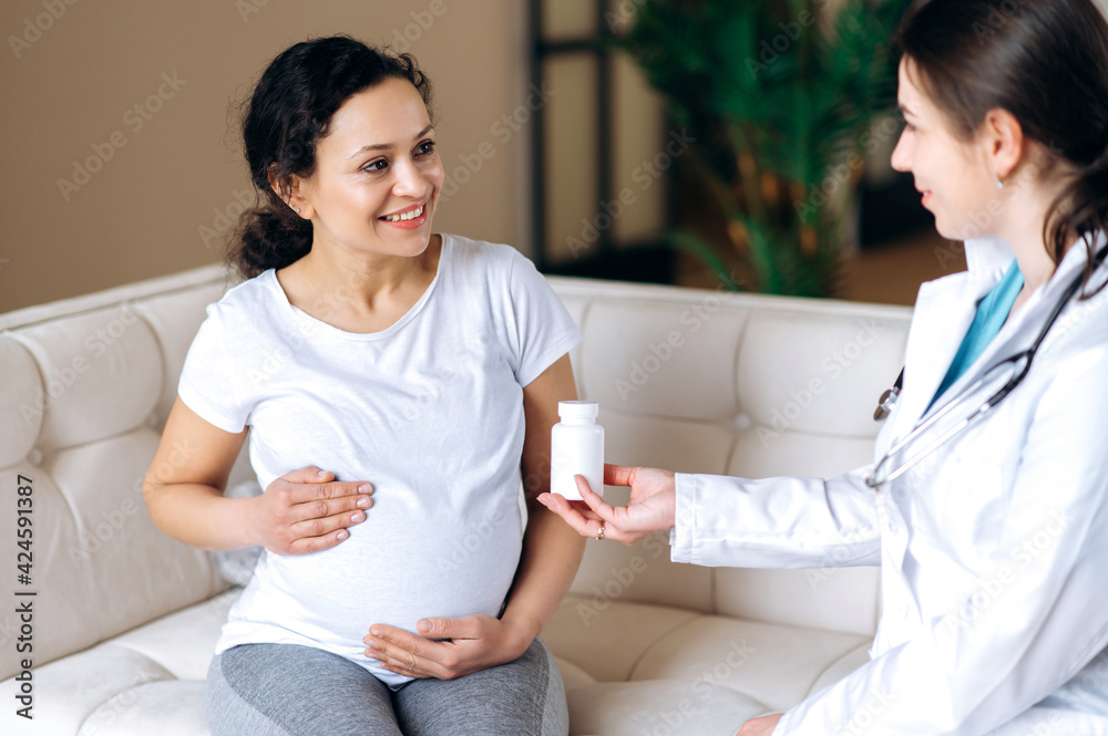 Fototapeta premium The doctor prescribes vitamins for a pregnant mixed race patient, sitting on the couch at home or in the hospital, the expectant mother stroking her belly. Healthy pregnancy concept