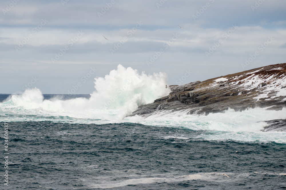 A rugged coastline on a sunny day with blue sky and clouds. The ocean is raging and is hitting the side of the craggy cliff. There's the land in the background and the teal ocean in the foreground.