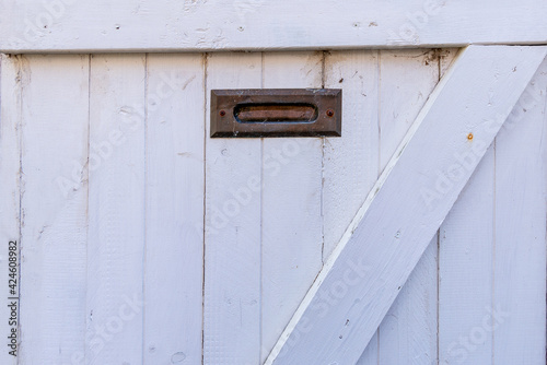A vintage exterior wooden white door with wood panels. There's a small brass post letter or mail slot at the top and center of the wood weather shutter door. The material is worn with specs of rust. © Dolores  Harvey