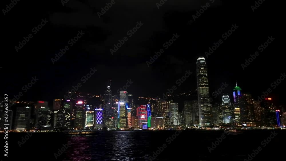 Wide view of famous Hong Kong skyline and Victoria Harbor at night with bright city lights, boats and ferries passing by on harbor. Hong Kong is a major modern financial hub in Asia