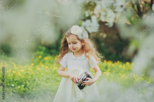 Little girl in white dress holding film camera in spring cherry garden. Portrait of happy child among white flowers trees. Childhood. Young photographer takes his first photos in sunny blooming park.