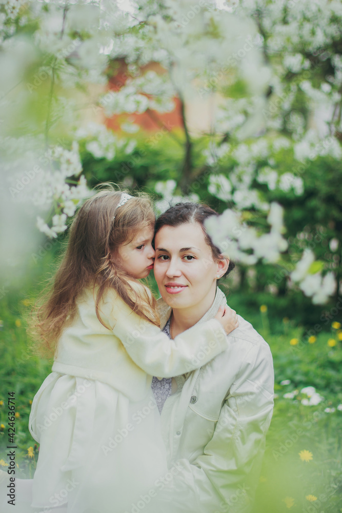 Happy mother's day. Little girl hugs her mother in the spring cherry garden. Portrait of happy mother and daughter among white flowers trees. Family values. Motherhood. Mom and child in blooming park. Happy mother's day. Little girl hugs her mother in the spring cherry garden. Portrait of happy mother and daughter among white flowers trees. Family values. Motherhood. Mom and child in blooming park.