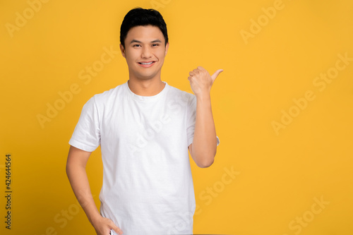 Young Asian man wearing a white T-shirt is pointing his finger with a happy face with copy space on a yellow background.