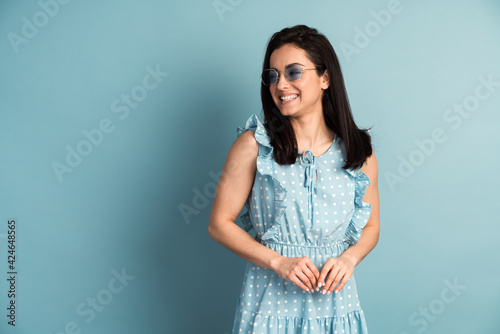 Charming, smiling brunette in a polka dot dress and sunglasses looks to the side. Beautiful girl on a background of a blue wall, opy space, place for text photo