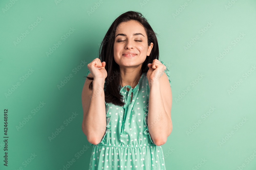 Amazing lady winning contest raising fists, wearing casual polka dot dress isolated on mint background. Girl gesturing on the background of an empty wall, copy space. Amazing lady winning contest raising fists, wearing casual polka dot dress isolated on mint background. Girl gesturing on the background of an empty wall, copy space.