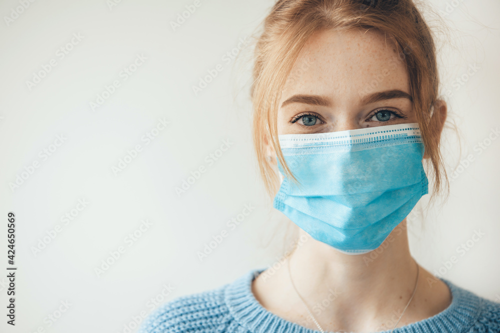 Close up photo of a caucasian woman with ginger hair and freckles smiling at camera wearing a medical mask near studio free space Close up photo of a caucasian woman with ginger hair and freckles smiling at camera wearing a medical mask near studio free space