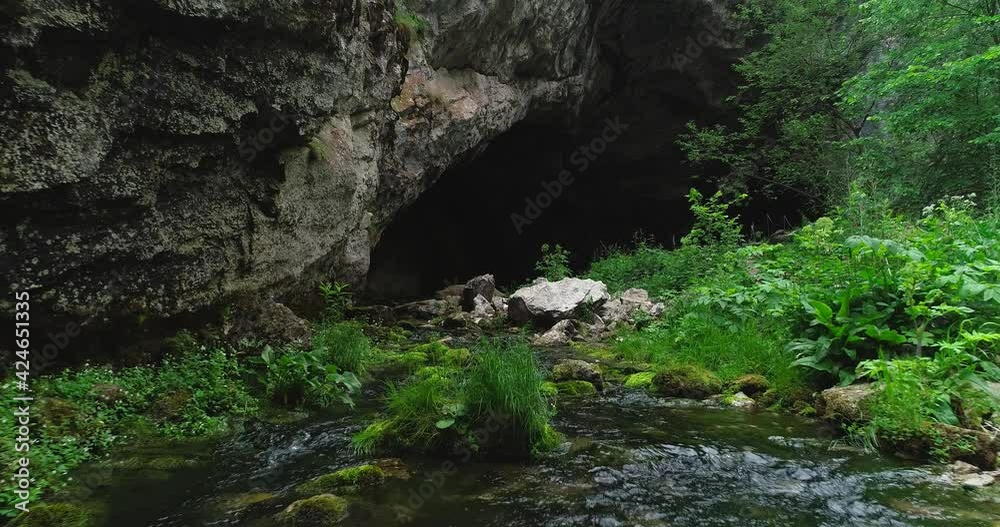 Kapova cave in Nature Reserve Shulgan-Tash. The entrance is huge arch with river flows out of ground. Limestone karst cave in the Burzyansky District of Bashkortostan. Aerial drone view at summer day Kapova cave in Nature Reserve Shulgan-Tash. The entrance is huge arch with river flows out of ground. Limestone karst cave in the Burzyansky District of Bashkortostan. Aerial drone view at summer day