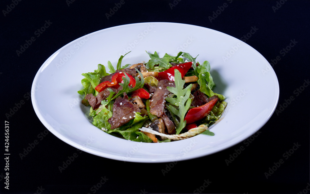 Salad with beef, arugula, red pepper, green lettuce, mushrooms in a white plate on a black background Salad with beef, arugula, red pepper, green lettuce, mushrooms in a white plate on a black background