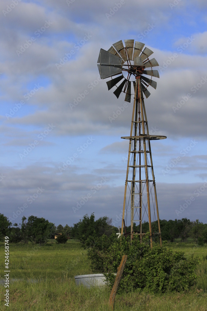 A Kansas Farm Windmill in a pasture with green grass, fence, tree,  with blue sky and clouds north of Hutchinson Kansas USA out in the country.