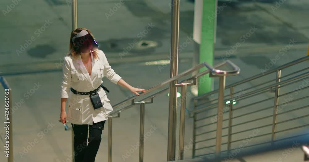Independent woman in fashion outfit and COVID face shield. Trendy looking blonde woman walking up by the stair at night city with steel and concrete architecture on background. Night scene footage Independent woman in fashion outfit and COVID face shield. Trendy looking blonde woman walking up by the stair at night city with steel and concrete architecture on background. Night scene footage
