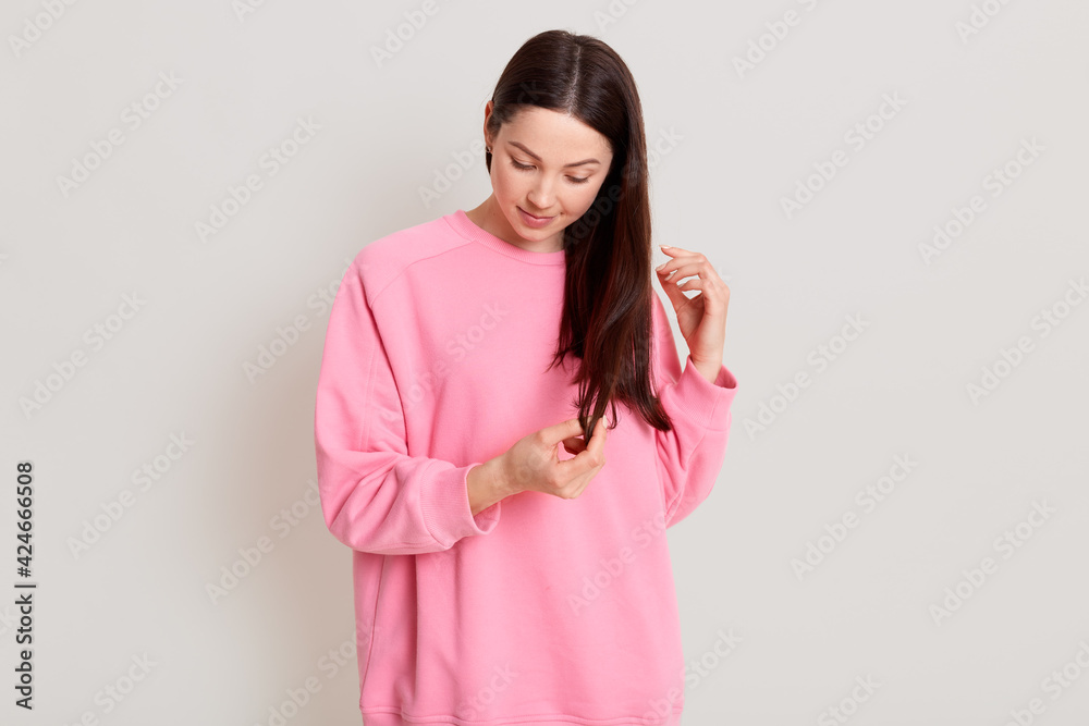 Horizontal shot of beautiful adorable young girl with dark haired wearing pink casual sweatshirt, standing isolated over white background, looking down and touching hair with hands. Horizontal shot of beautiful adorable young girl with dark haired wearing pink casual sweatshirt, standing isolated over white background, looking down and touching hair with hands.