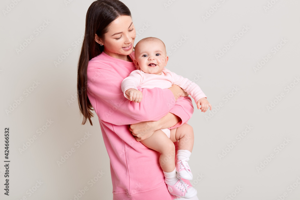 Dark haired young mother hugging her daughter, baby smiling, mommy looking at infant, wearing casual pink sweater and white pants, isolated over light background. Dark haired young mother hugging her daughter, baby smiling, mommy looking at infant, wearing casual pink sweater and white pants, isolated over light background.
